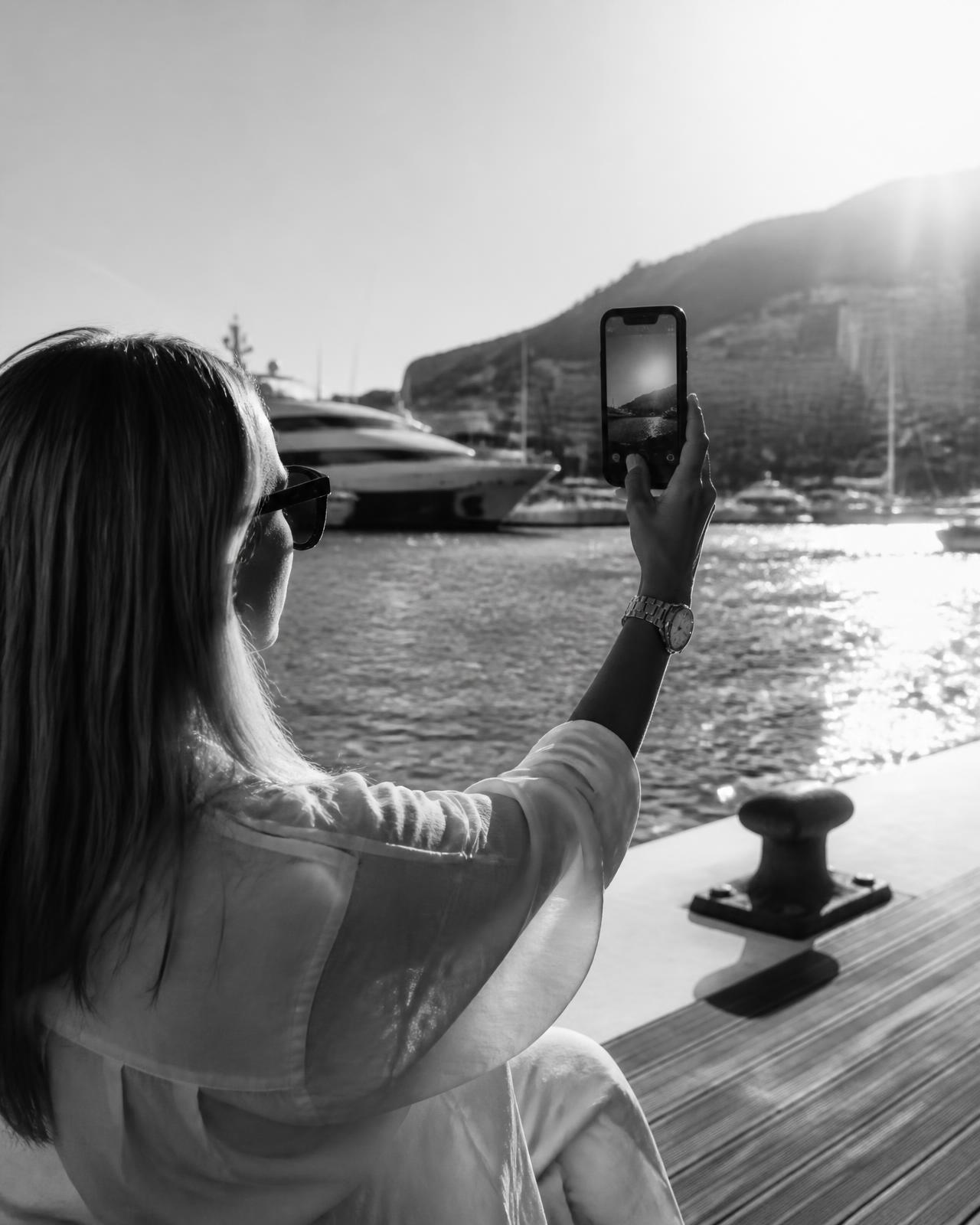 A visitor holding a phone vertically toward the sea at a luxury yacht marina, viewing a yacht in augmented reality